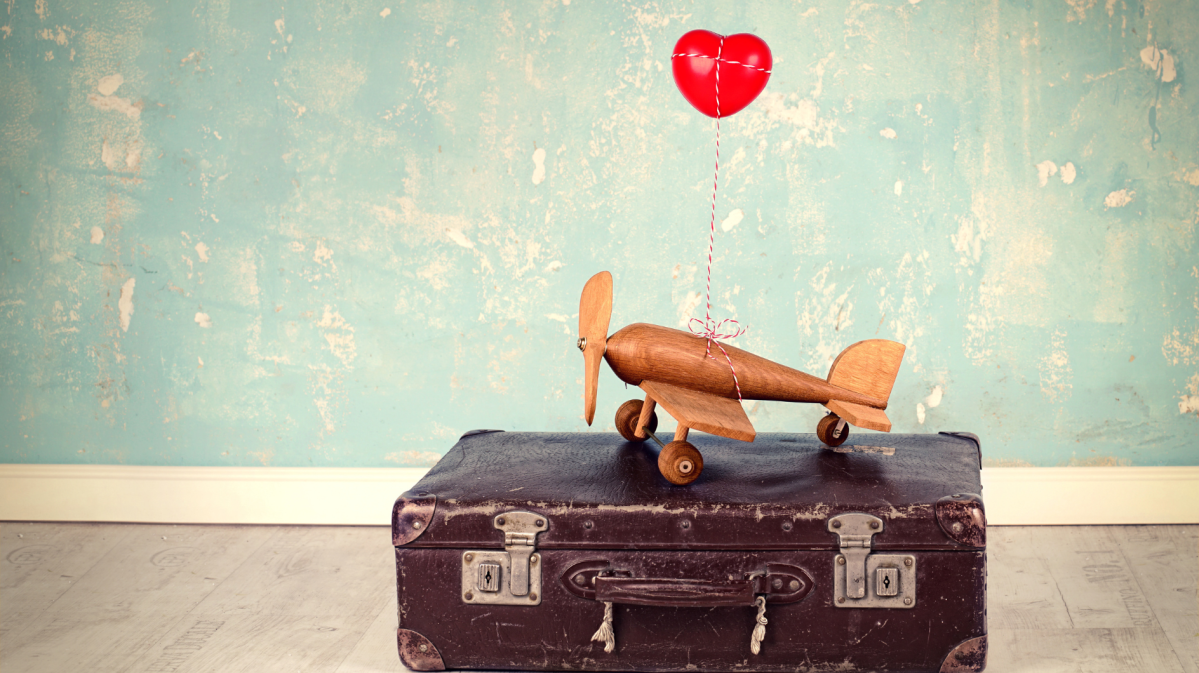 Auf einem alten brauen Koffer stejt ein kleines gelbes Flugzeug mit einem roten Herzluftballon angehängt.