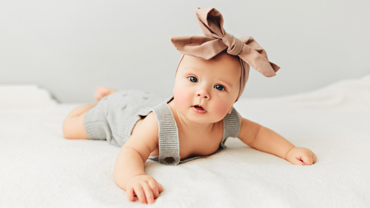 Süßes Baby liegt auf dem Bett und lächelt. Es trägt eine Schleife auf dem Kopf.