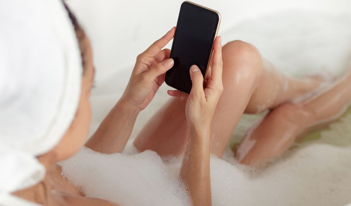 Frau liegt in der Badewanne, mit Smartphone in der Hand