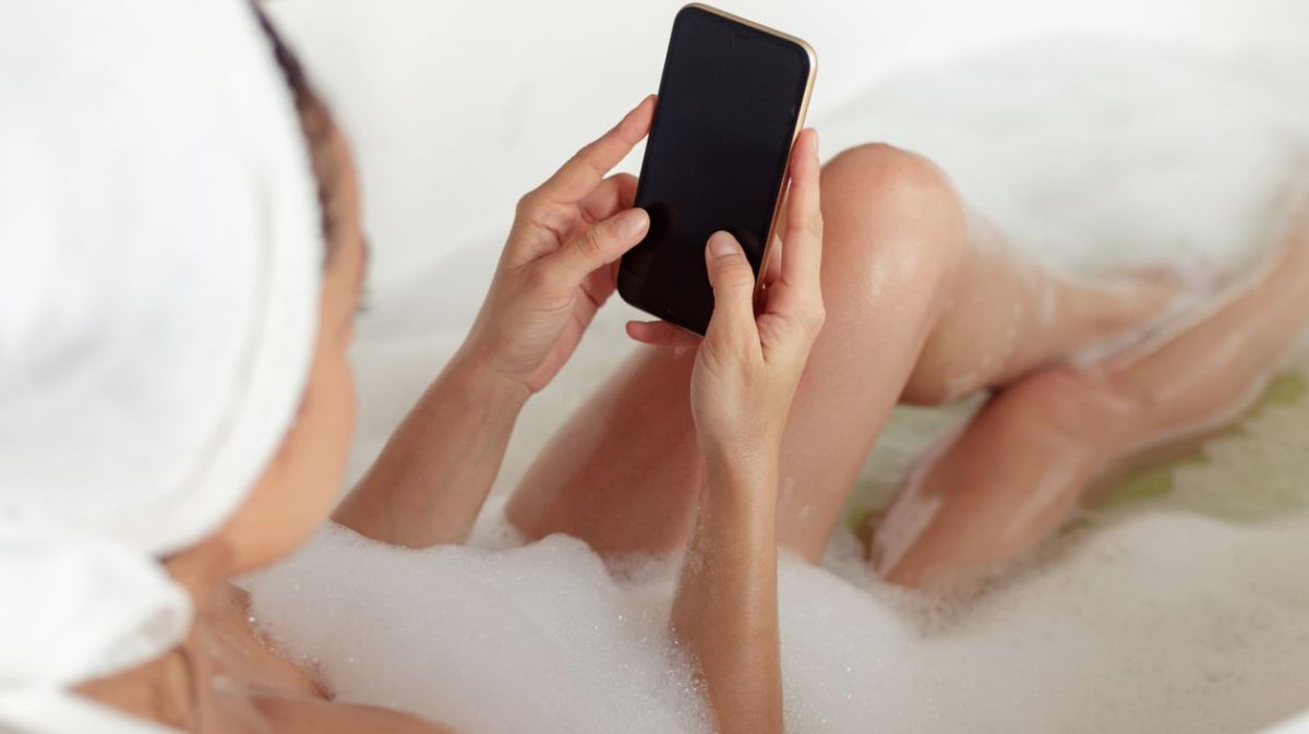 Frau liegt in der Badewanne, mit Smartphone in der Hand