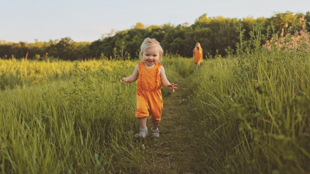 Kleines Mädchen läuft fröhlich über eine Wiese, im Hintergrund steht die Mutter.