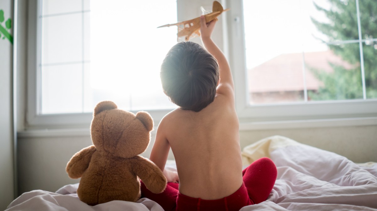 Fröhliches Kind, das mit Holzspielzeugflugzeug und Teddybär im Bett spielt