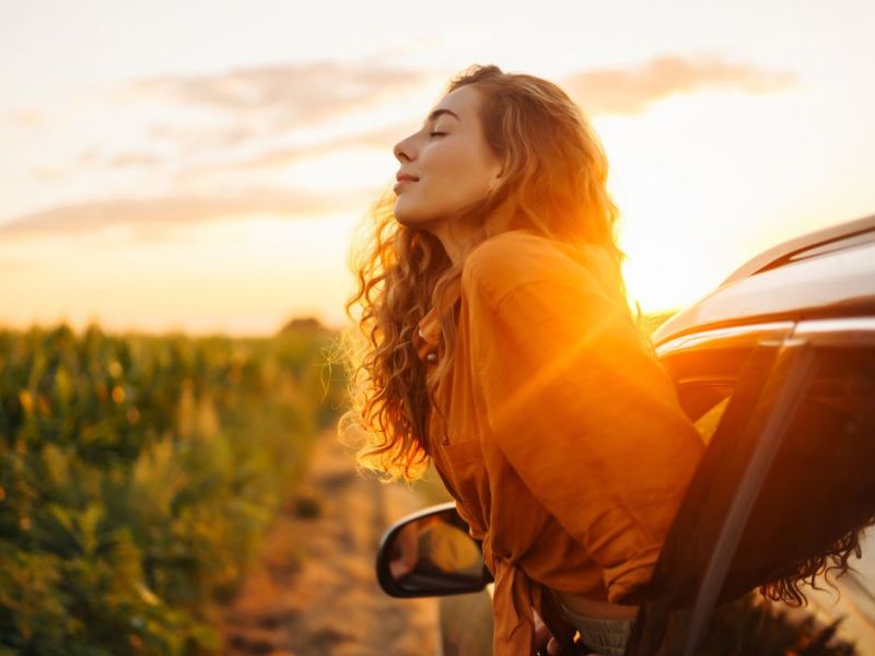 Frau, die sich aus dem Fenster im Auto lehnt und die Sonnenstrahlen genießt