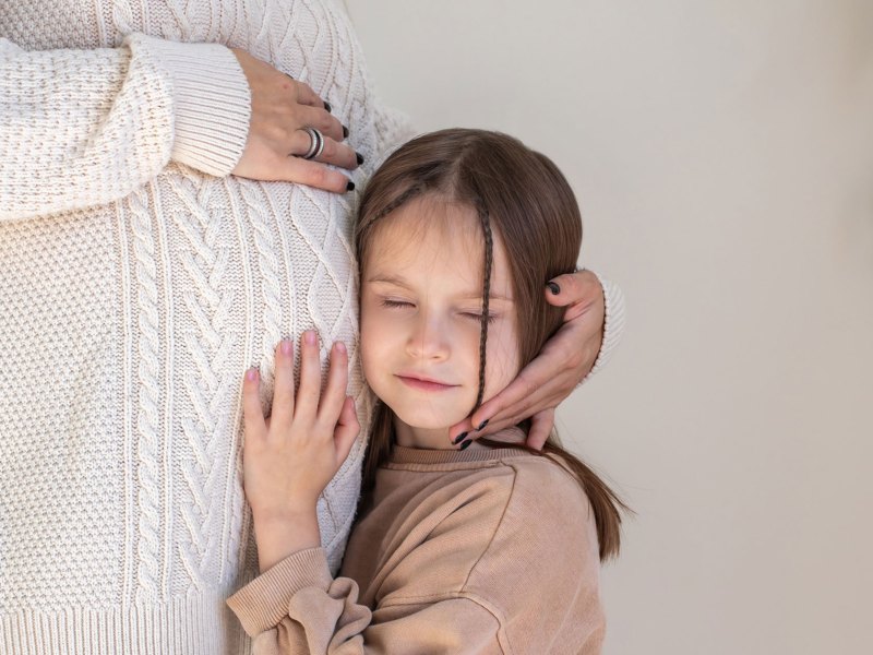 Kind hält sein Ohr an den Bauch einer Schwangeren