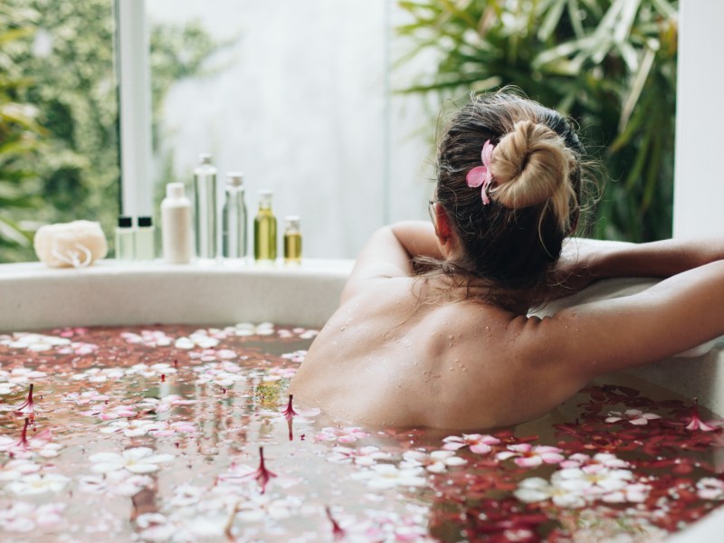 Frau in einem Bad voller Rosenblüten, die ihre Haare hochgesteckt hat und nach draußen zum Fenster blickt