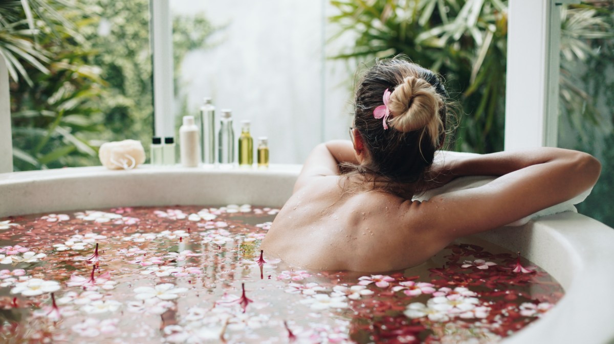 Frau in einem Bad voller Rosenblüten, die ihre Haare hochgesteckt hat und nach draußen zum Fenster blickt