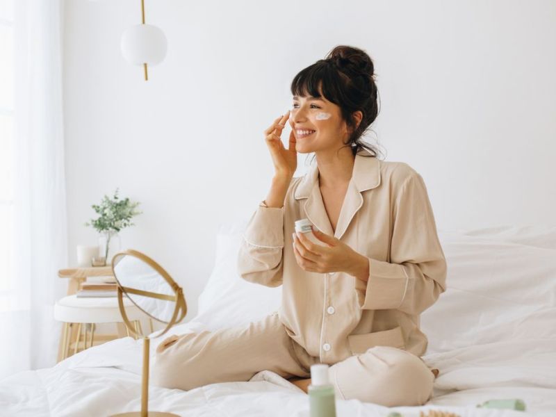 Frau sitzt mit Gesichtscreme im Bett und lacht