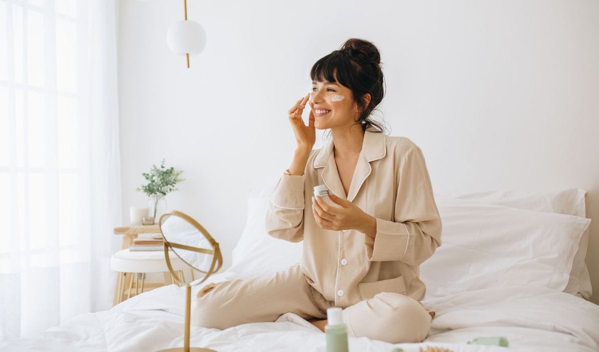 Frau sitzt mit Gesichtscreme im Bett und lacht