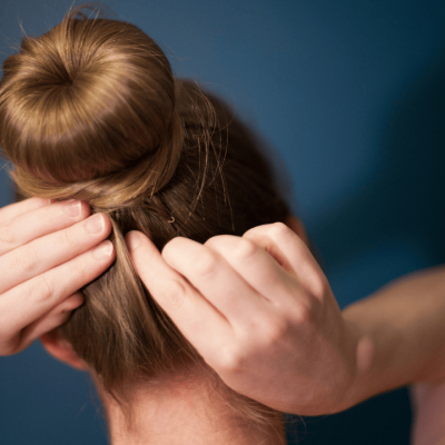 Eine Frau, die sich von hinten gesehen die Haare zu einem Knoten zusammenbindet. Sie steckt Haarklammern, Bobby Pins, in den Knoten, um ihn am Kopf zu befestigen.