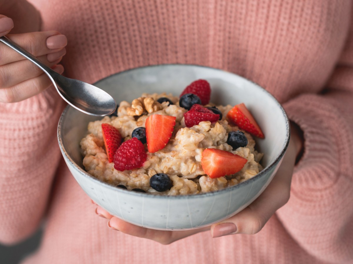 Frau mit rosa Pullover h&auml;lt Sch&uuml;ssel mit Porridge und Fr&uuml;chten in Hand.