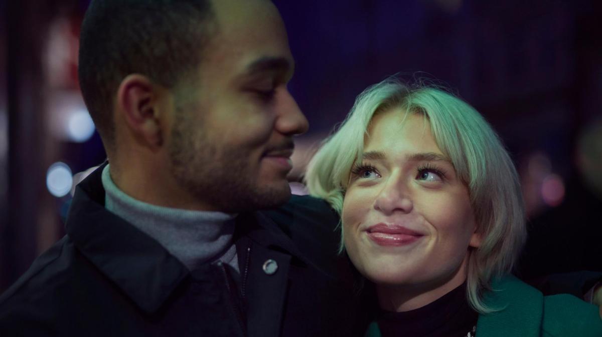 Ein junges Paar blickt sich lächelnd an, die Frau mit blondem Haar schaut verliebt hoch, während der Mann sanft zurücklächelt.