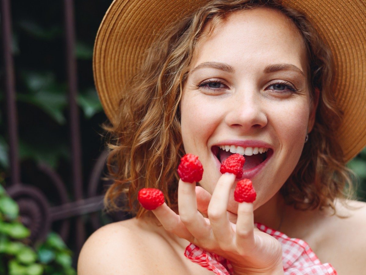 Frau mit Hut, hat sich Himbeeren auf die Fingerspitzen gesteckt.