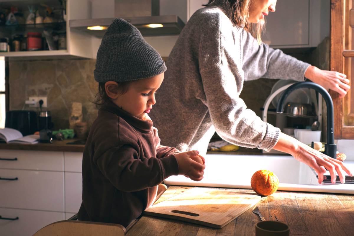 Kleiner Junge steht konzentriert an der Küchenzeile und schält sich allein eine Orange. Die Mutter wäscht daneben ab.