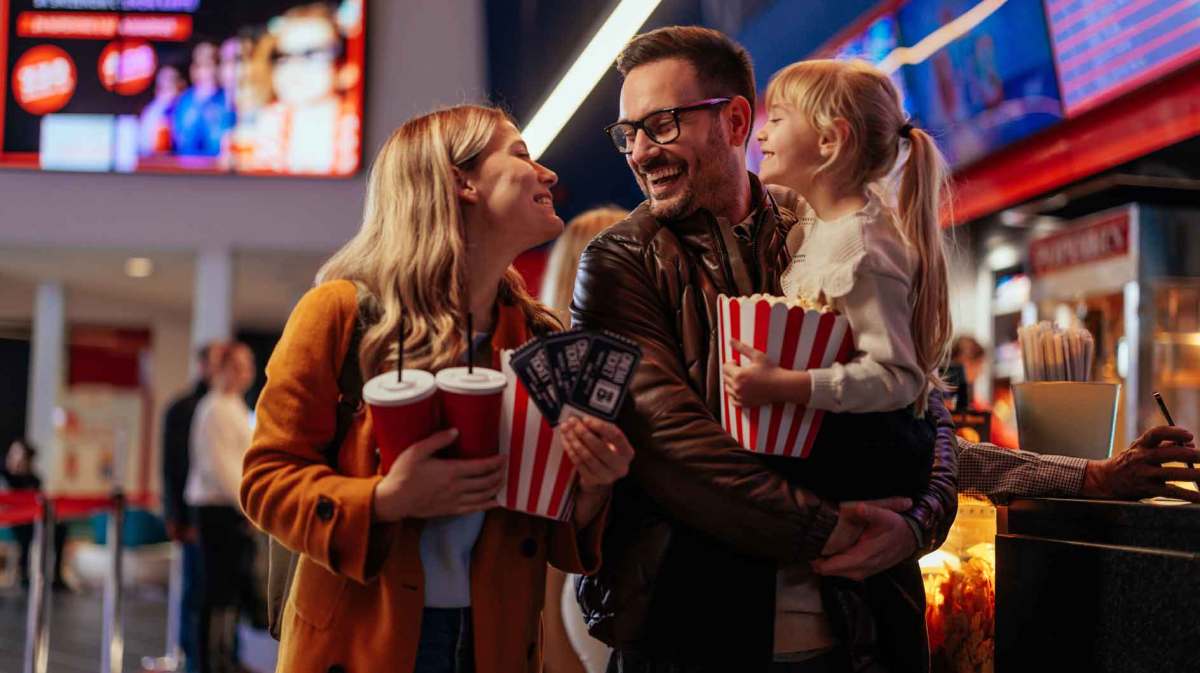 Eltern und Tochter im Kino. Vater trägt seine Tochter, Mutter und Kind halten eine Portion Popcorn in den Armen.