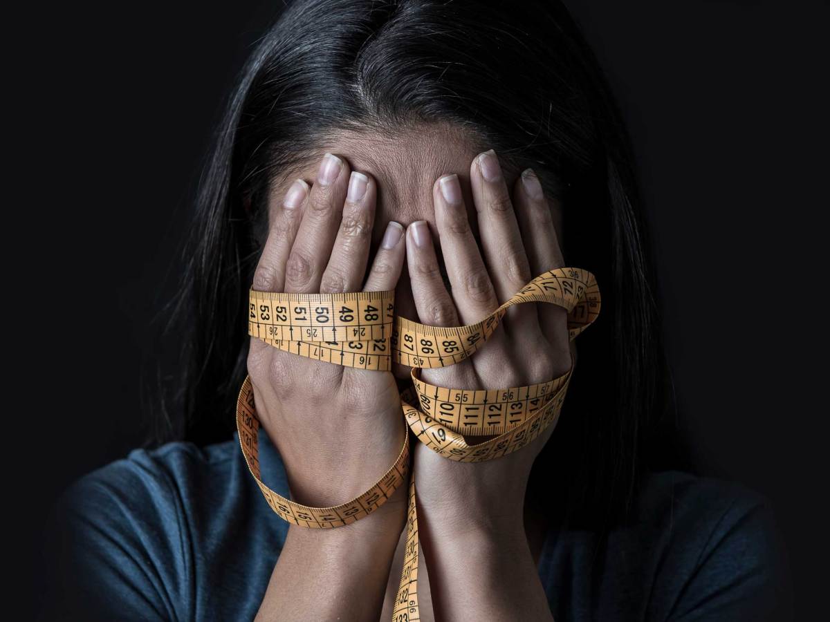 mit Maßband umwickelte Hände, die das Gesicht eines jungen, deprimierten und besorgten Mädchens bedecken, das an Magersucht oder Bulimie leidet