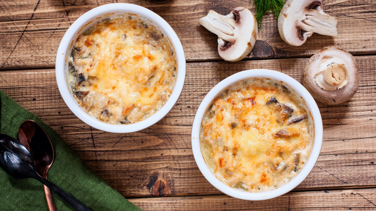 Draufsicht: Zwei runde Auflaufformen einem Penne-Champignon-Auflauf stehen auf einem Holztisch. Drum herum liegen frische Pilze.