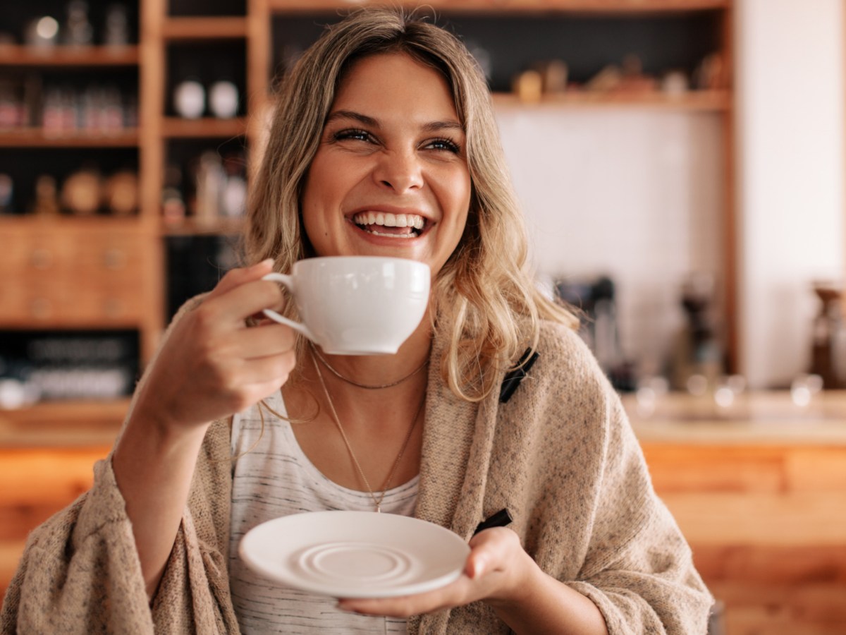 Frau mit blonden Haaren lacht und h&auml;lt Kaffeetasse in Hand.