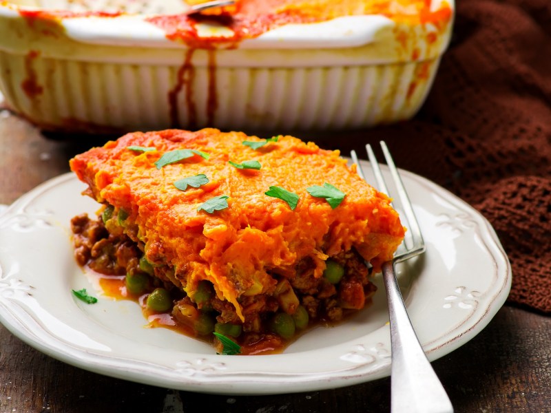 Ein Stück Shepherd's Pie mit Süßkartoffelbrei auf einem Teller, daneben eine Gabel, dahinter die Auflaufform.