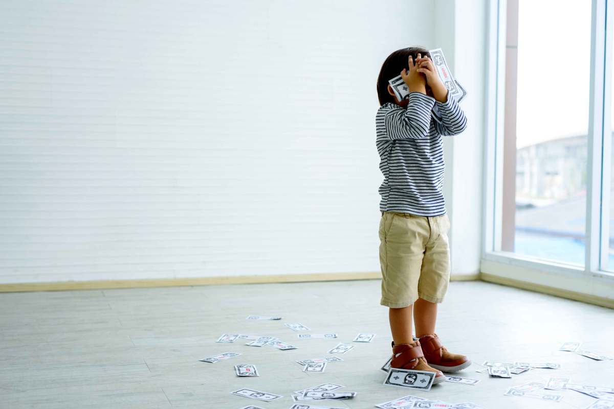 Kleiner Junge steht in einem leeren Zimmer und hält sich seine Hände, die voll mit Geldscheinen sind, vor das Gesicht. Um ihn herum liegen weitere Scheine auf dem Boden.