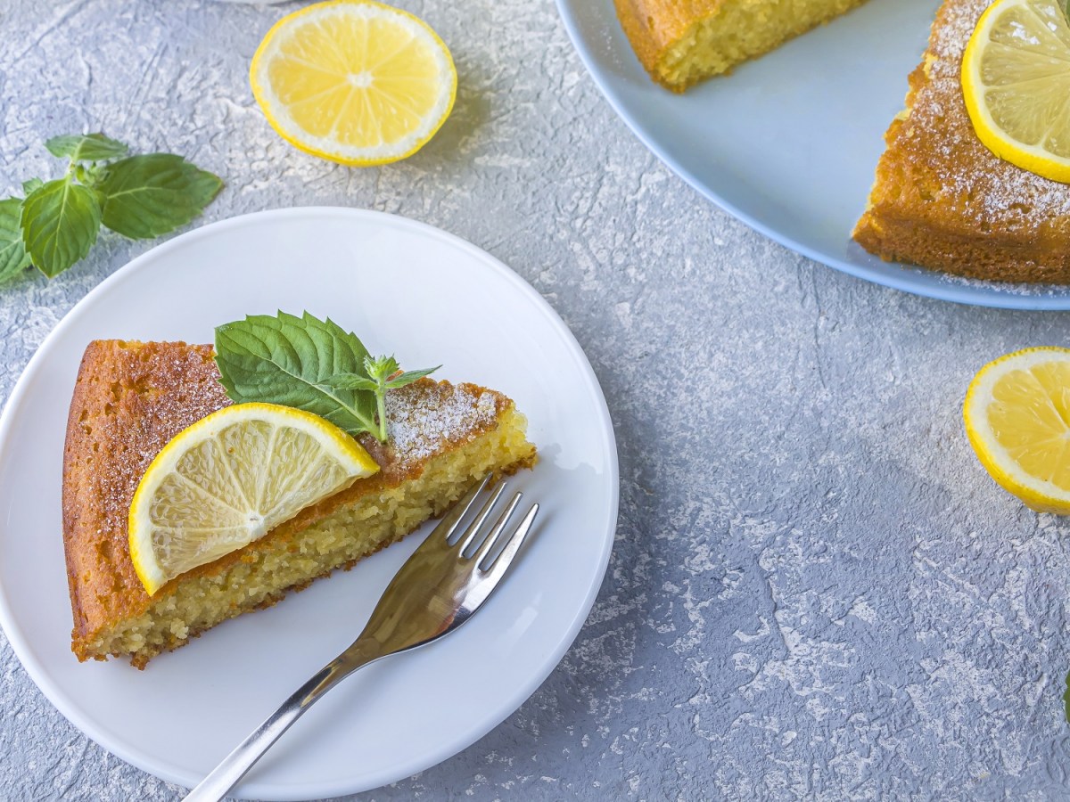 Draufsicht: ein angeschnittener Zitronen-Grießkuchen. Einige Kuchenstücke liegen auf zwei blauen Tellern verteilt. Die Kuchen sind mit Zitronenscheiben und Minzblättern garniert.