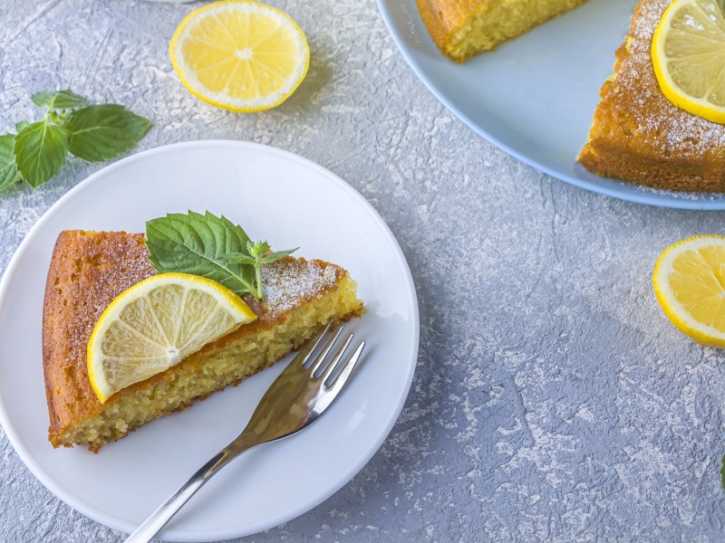 Draufsicht: ein angeschnittener Zitronen-Grießkuchen. Einige Kuchenstücke liegen auf zwei blauen Tellern verteilt. Die Kuchen sind mit Zitronenscheiben und Minzblättern garniert.