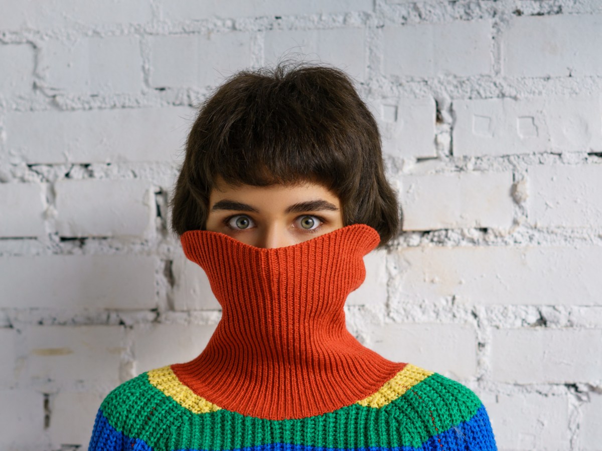 Frau mit buntem Pulli vor Wand, die den Rollkragen &uuml;ber Nase und Mund zieht