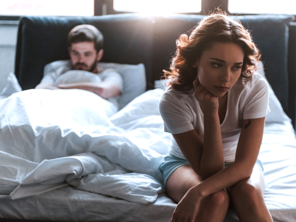 Frau auf der Bettkante einer Matratze, die besorgt nach vorne blickt, w&auml;hrend im Hintergrund ein Mann auf der Matratze liegt.