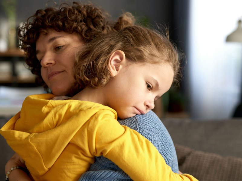 Mutter und Tochter umarmen sich. Das Mädchen, ca. 10 Jahre, sieht traurig und enttäuscht aus.