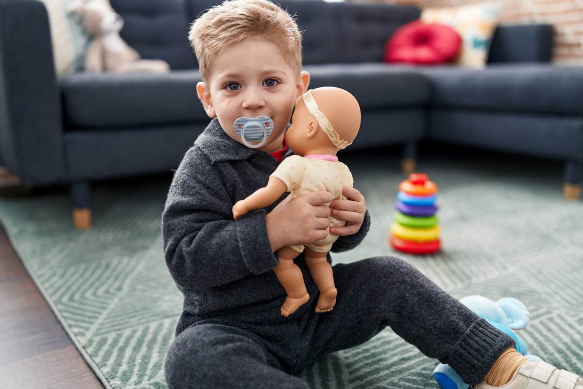 Ca. dreijähriger Junge sitzt mit einer Puppe im Arm und einem Nuckel im Mund im Zimmer und spielt.