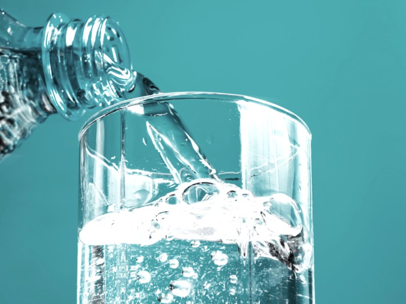 Mineralwasser wird aus einer Glasflasche in ein Glas geschüttet. Der Hintergrund ist türkis.