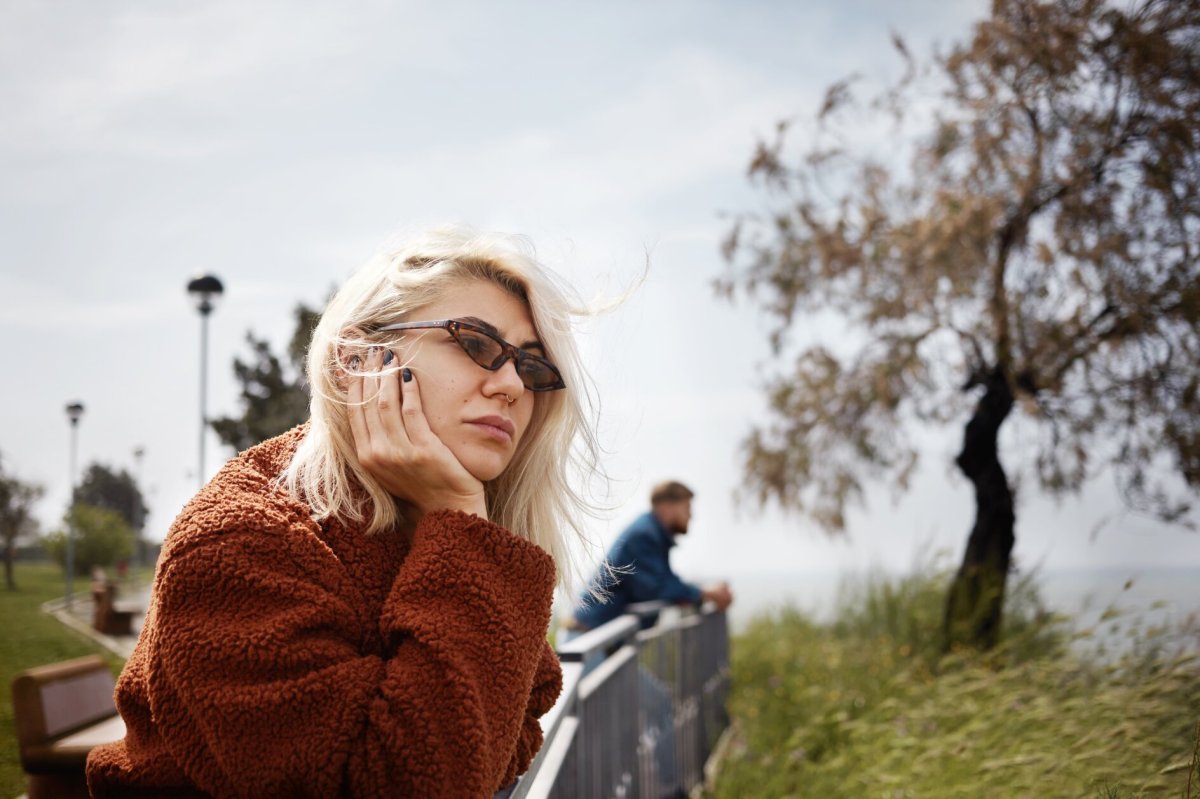 Frau schaut mit Sonnenbrille gelangweilt von einer Brücke hinunter, im Hintergrund ein Mann der ebenfalls herunterschaut