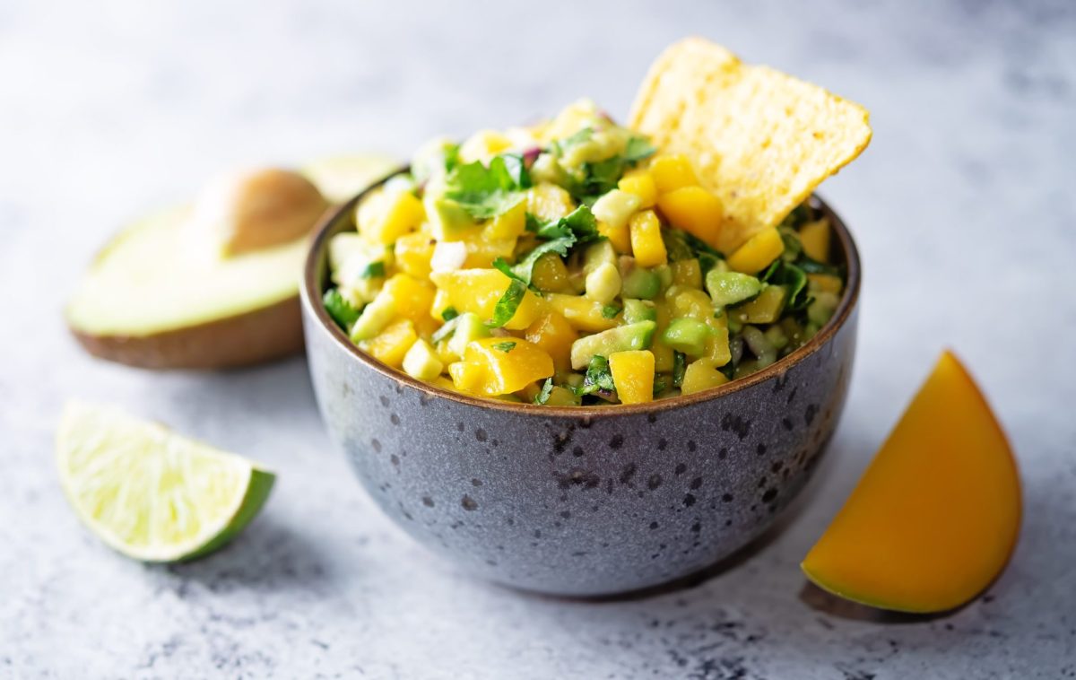 Eine Schüssel Mango-Avocado-Salsa mit eine Nacho-Chip, daneben je eine angeschnittene Limette, Avocado und Mango.