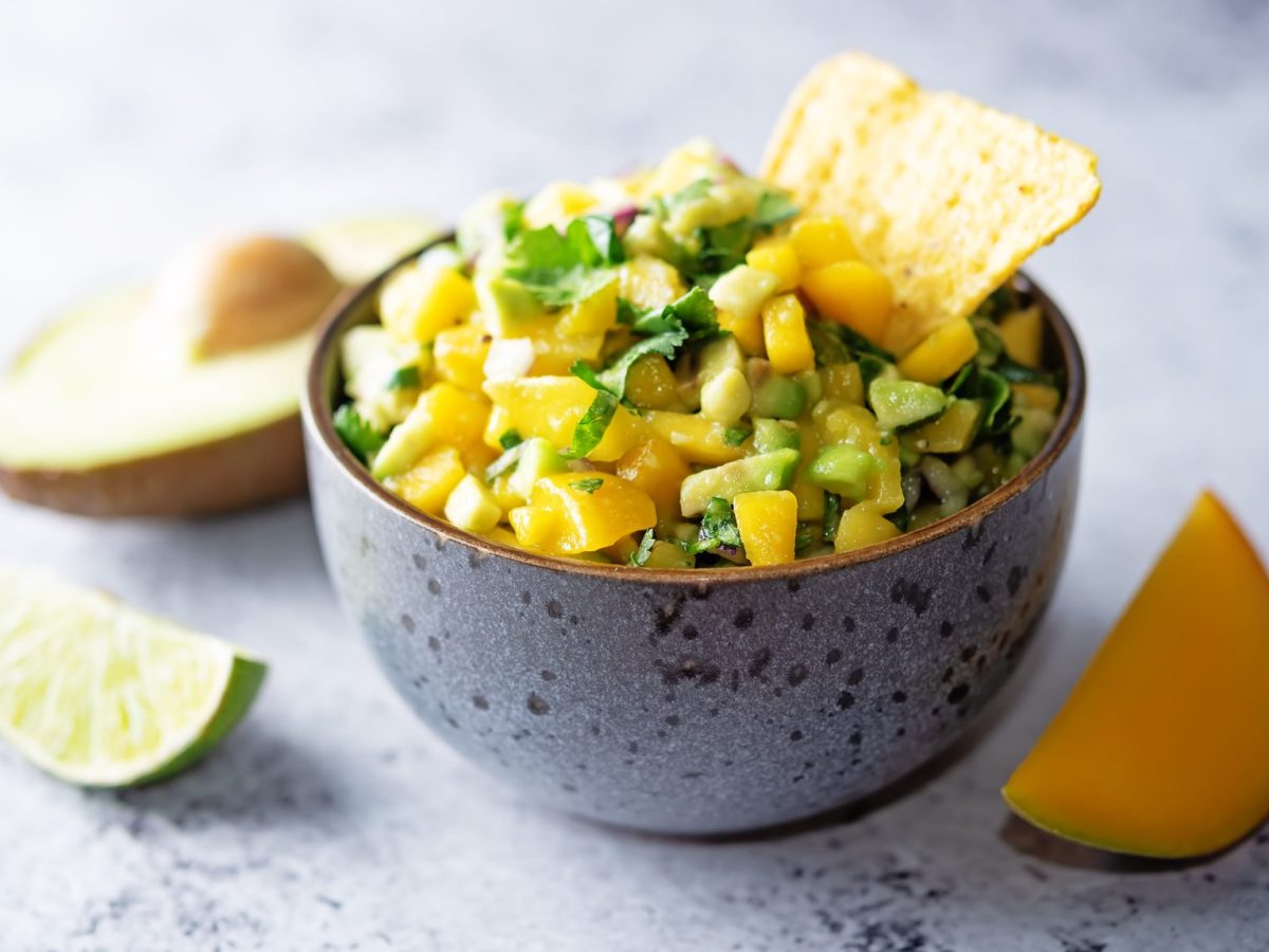 Eine Schüssel Mango-Avocado-Salsa mit eine Nacho-Chip, daneben je eine angeschnittene Limette, Avocado und Mango.