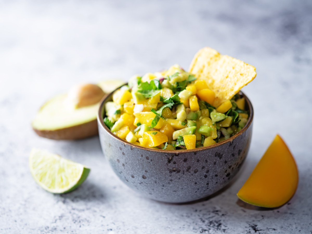 Eine Schüssel Mango-Avocado-Salsa mit eine Nacho-Chip, daneben je eine angeschnittene Limette, Avocado und Mango.