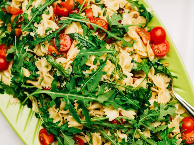 Nudelsalat mit Farfalle, Rucola und Tomaten in einer grünen Servierschüssel.