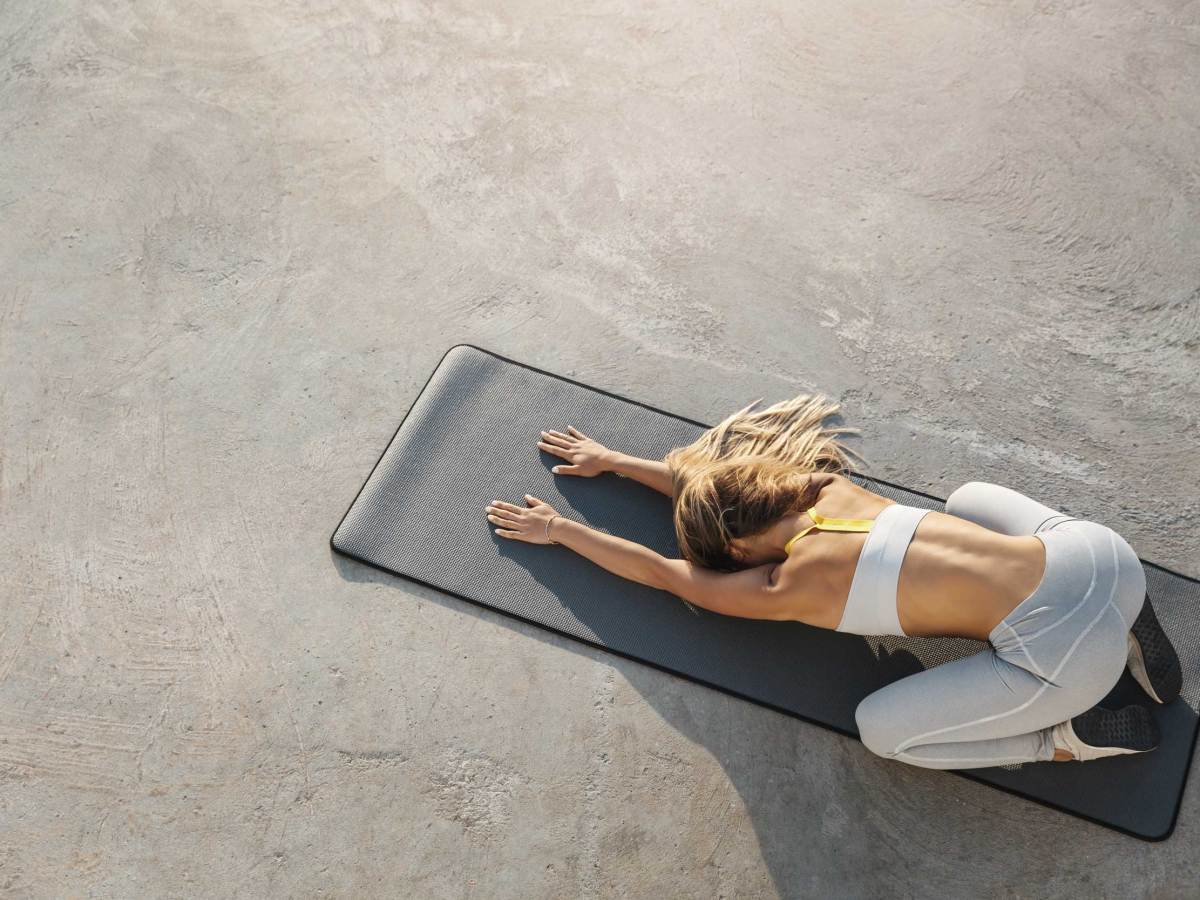 Attraktive, sportliche Frau, die auf einer Yogamatte sitzt und Balasana, die Kinderhaltung, dehnt, trainiert, Asana-Posen während des morgendlichen Trainings macht, die Hände nach vorne ausstreckt