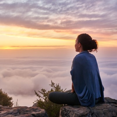 Frau auf Berg, die auf einem Felsen im Schneidersitz sitzt und über den Wolken den Sonnenaufgang bestaunt