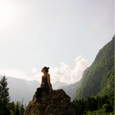 Frau auf einem Felsen in einer Bergregion, die nach oben in den Himmel schaut