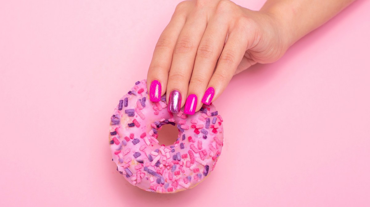 Pinke Nägel, die einen pinken Donut in der Hand halten und vor einer rosa Wand posieren