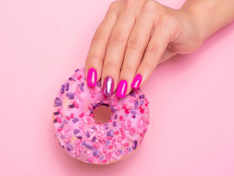 Pinke Nägel, die einen pinken Donut in der Hand halten und vor einer rosa Wand posieren