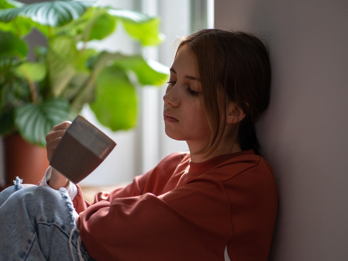 Frau auf Boden mit Kaffetasse in der Hand, die sich erschöpft gegen die Wand lehnt, mit leerem Blick