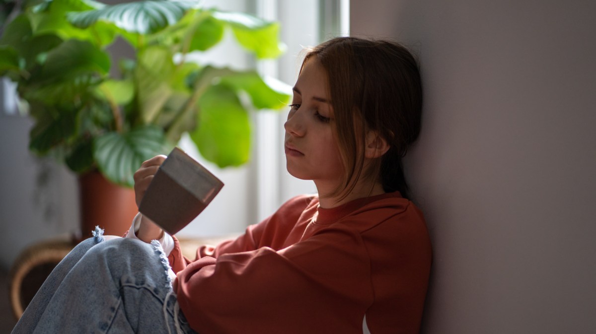 Frau auf Boden mit Kaffetasse in der Hand, die sich erschöpft gegen die Wand lehnt, mit leerem Blick