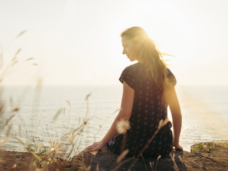 Frau an Klippe am Meer, die durch die Sonne nach hinten in die Kamera blickt