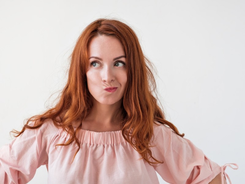 Frau mit roten Haaren guckt mit Schnute nach links vor weißer Wand