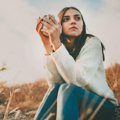 Frau mit Kaffeetasse in der Hand, die in einem Feld sitzt und zur Seite schaut