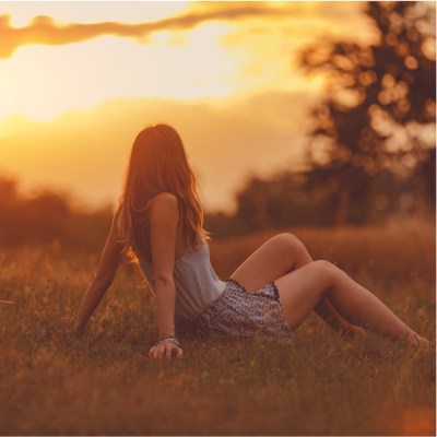 Frau im Sommer auf einem Feld, die sitzt und sich mit den Händen abstützt, während sie den Sonnenuntergang anschaut