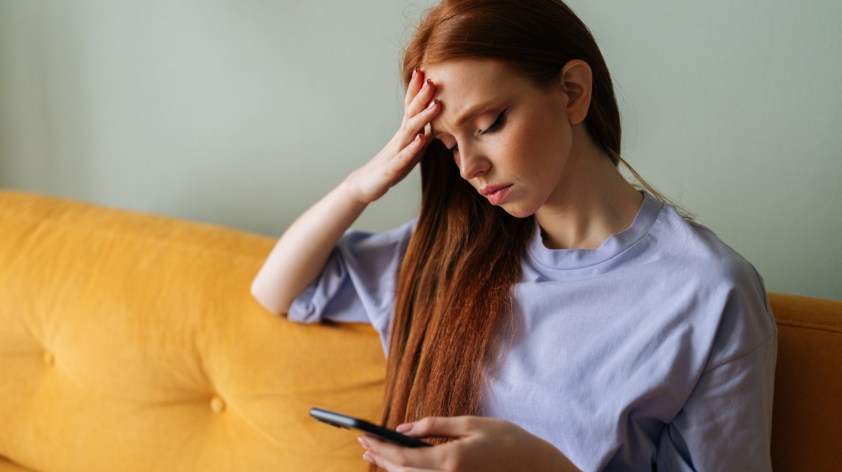 Rothaarige Frau auf gelber Couch, die nachdenklich auf Handy schaut