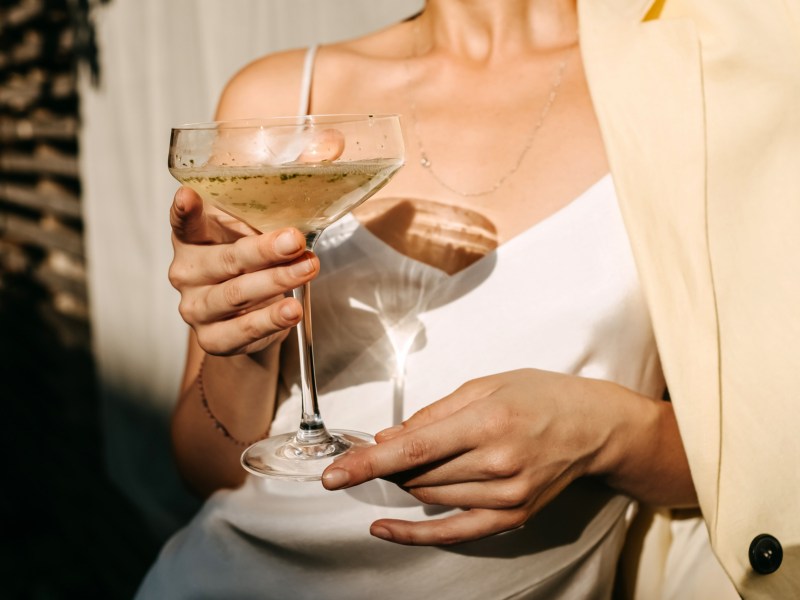 Frau in elegantem weißen Kleid, die ein Cocktailglas in der Hand hält und von der Sonne bestrahlt wird