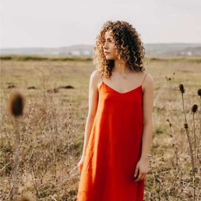 Frau in rotem, langen Kleid am Strand, die mit ihren lockigen, braunen Haaren zur Seite blickt