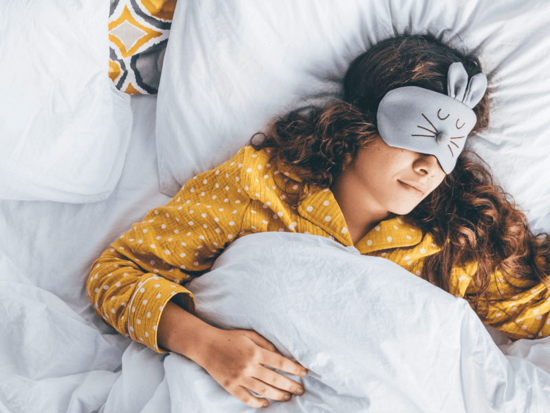 Millennial-Frau im Bett liegend mit Schlafmaske und Telefon in der Hand.